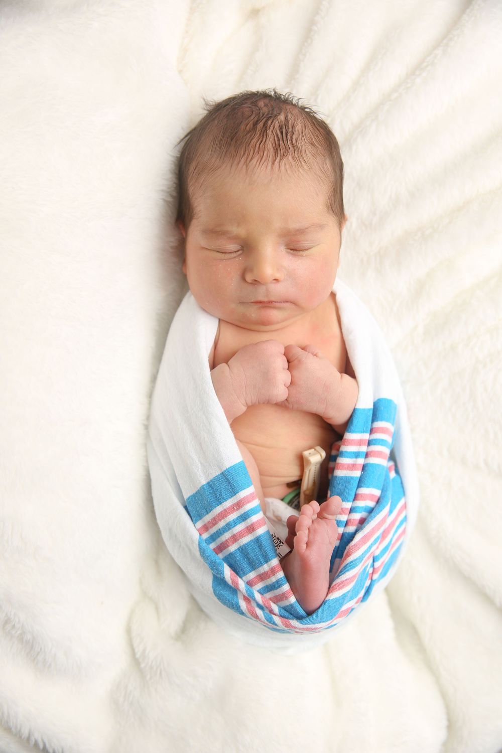 Peaceful newborn baby sleeping swaddled in a striped hospital blanket on a soft white surface.