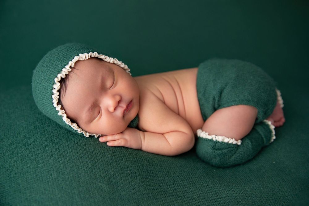 Newborn baby sleeping peacefully in green knit outfit and bonnet on a green backdrop.
