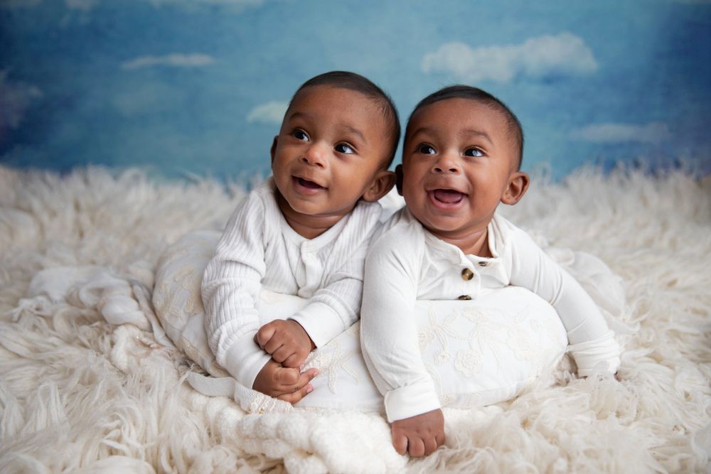 Two adorable Black twin babies in white onesies smiling on a fluffy white blanket with a blue sky backdrop.