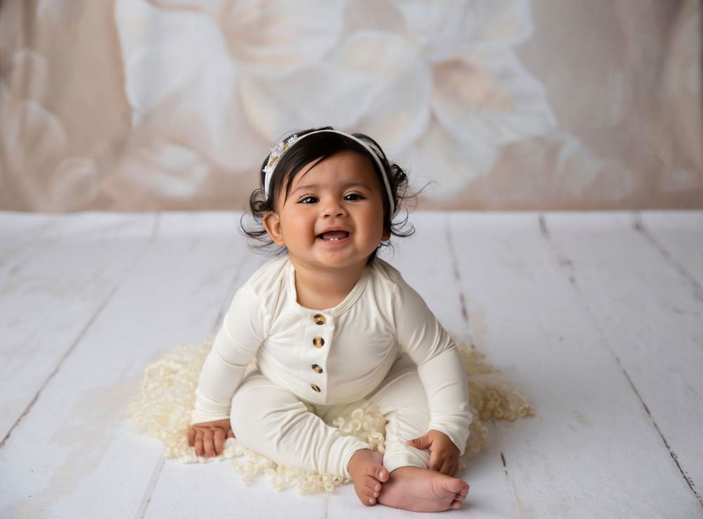 Smiling baby in cream romper sits on white wood floor against a soft floral backdrop.