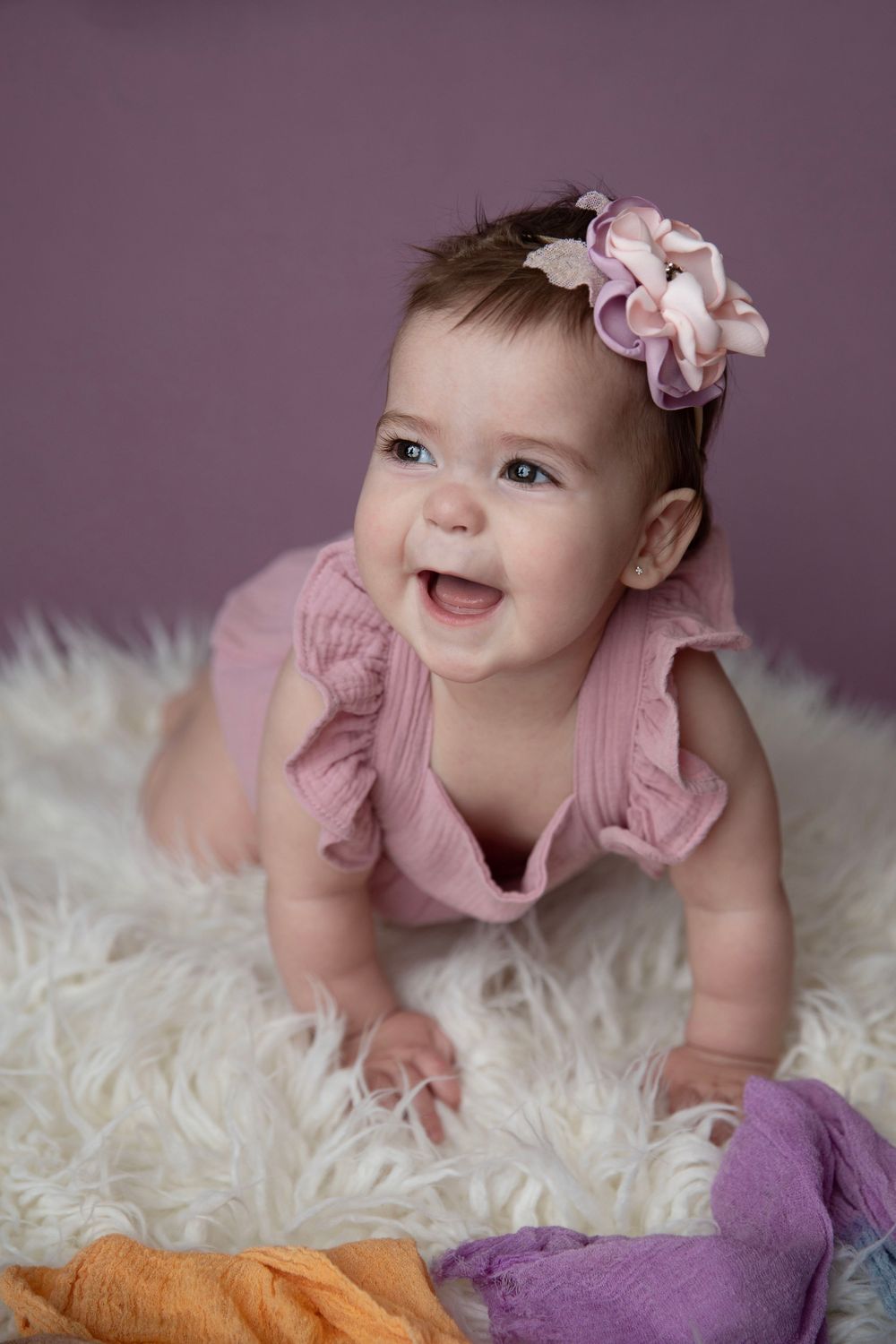Smiling baby girl in pink romper and floral headband crawling on white fluffy rug with colorful blankets.