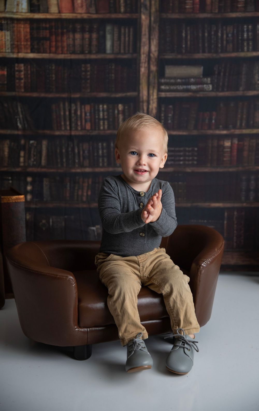 Smiling toddler boy sitting in a brown leather mini chair with a library bookshelf backdrop, clapping hands.
