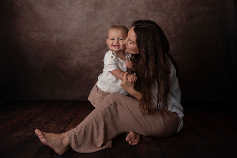 A mother sits on a wooden floor kissing her smiling baby dressed in white against a brown backdrop.