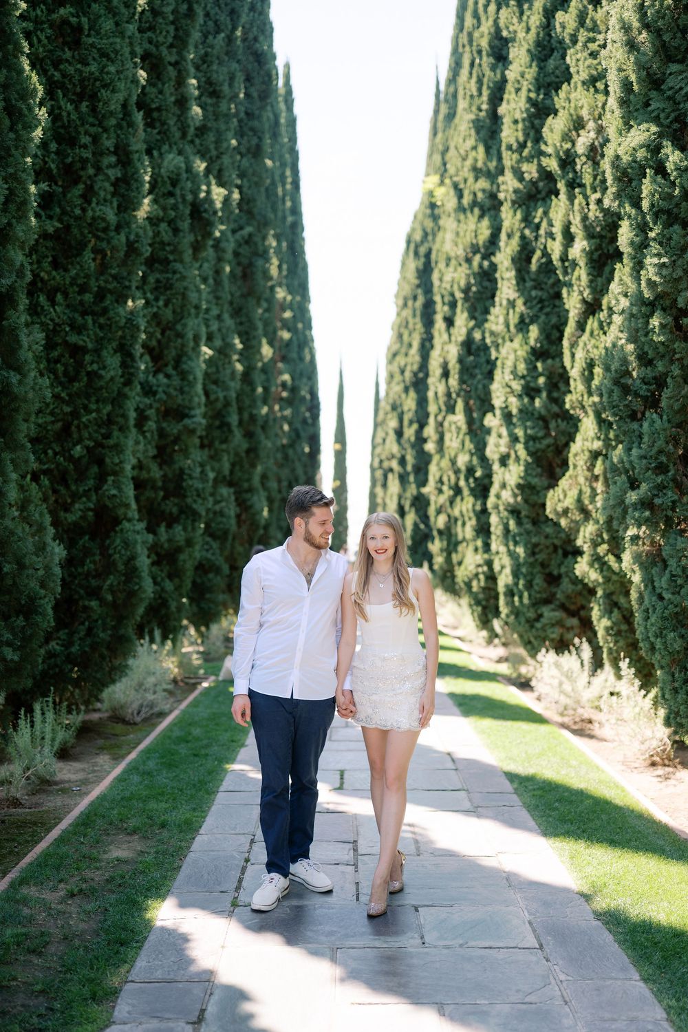 Couple walking hand in hand along a stone path lined with tall cypress trees in a formal garden.
