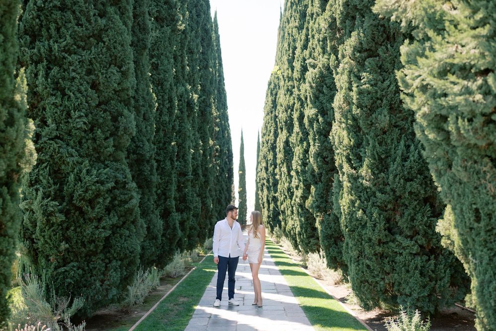 Couple walking hand in hand along a cypress tree-lined pathway at a Tuscan villa on a sunny day.