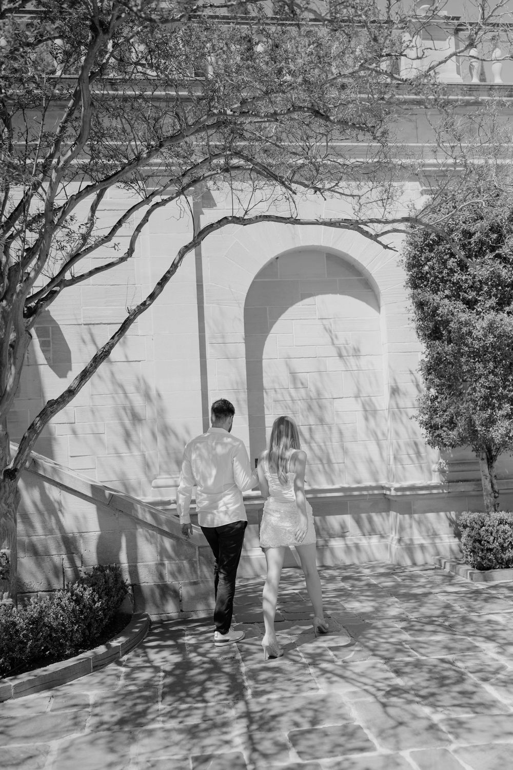 Black and white photo of a couple walking hand in hand toward a sunlit arched wall amid tree shadows.