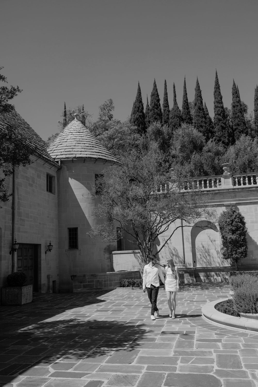 Black and white photo of a couple walking hand in hand through an elegant stone courtyard with cypress trees.