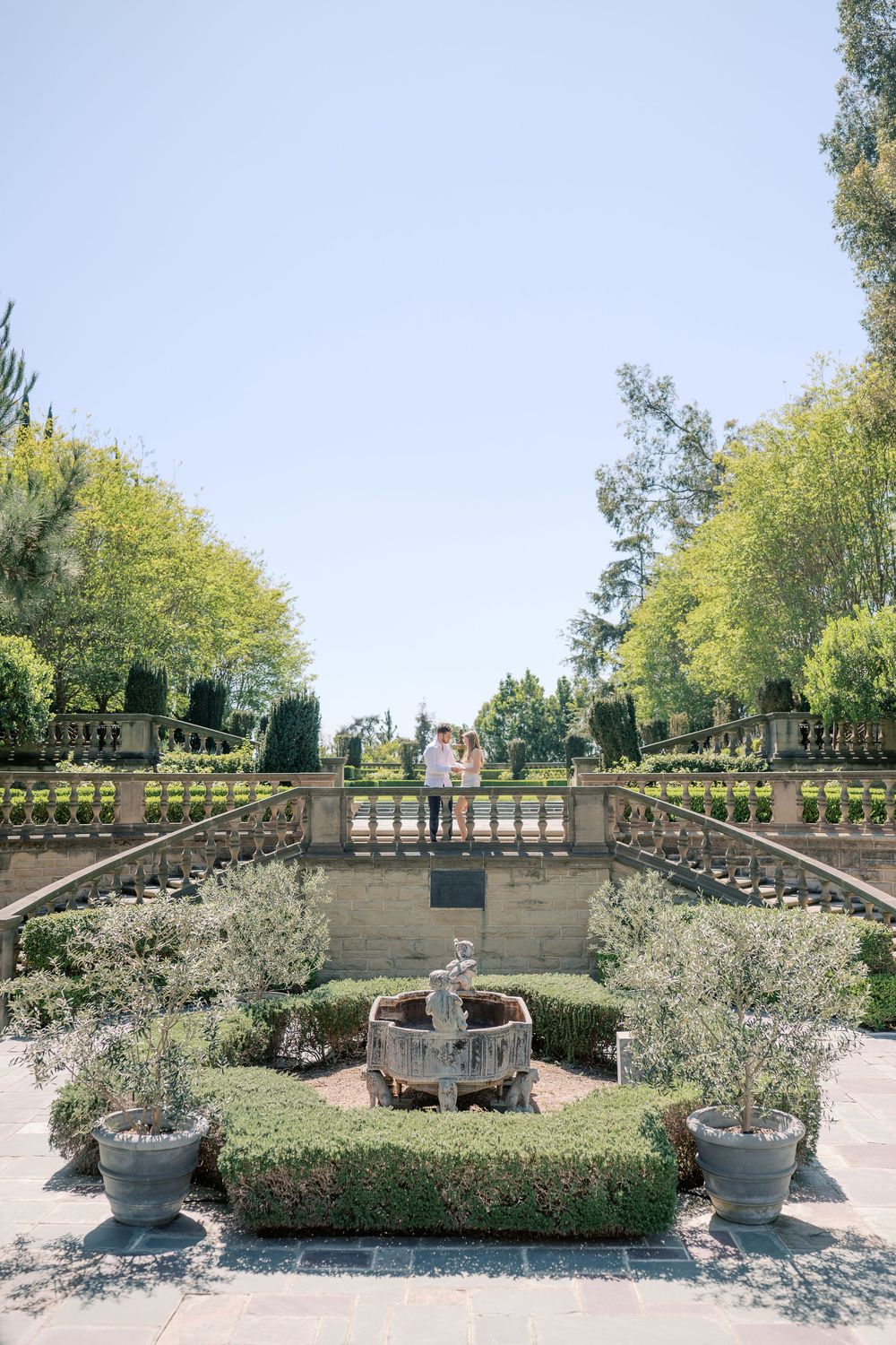 Couple standing on a stone balustrade terrace overlooking a formal garden with fountain and manicured hedges.