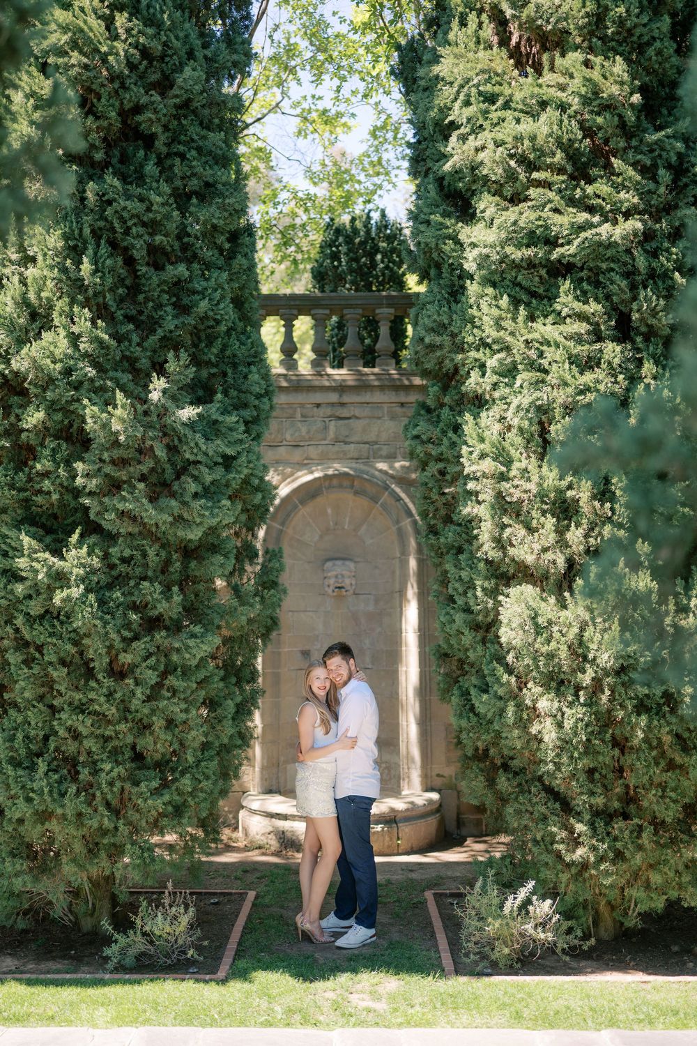 Couple embracing in a romantic Italian-style garden niche framed by tall cypress trees and stone architecture.