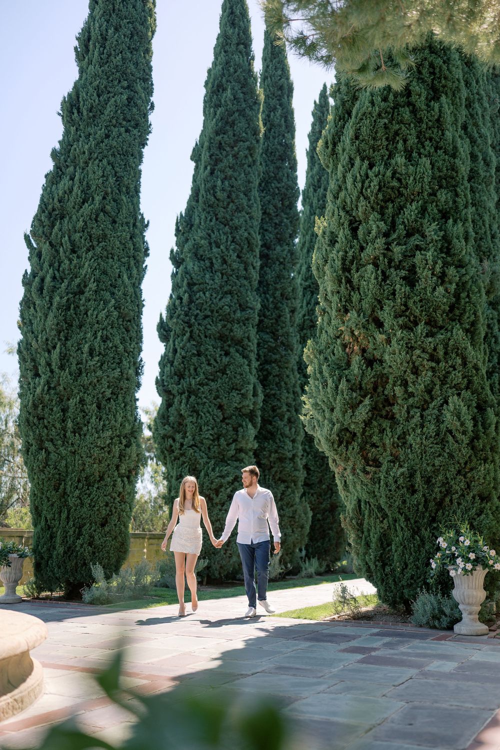 Couple holding hands walking along a garden path lined with tall cypress trees on a sunny day.