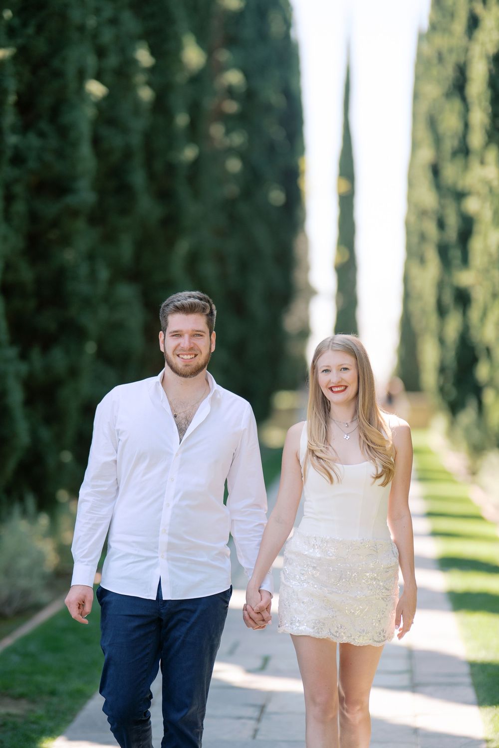 Happy couple holding hands walking along a tree-lined path, man in white shirt and woman in white mini dress.
