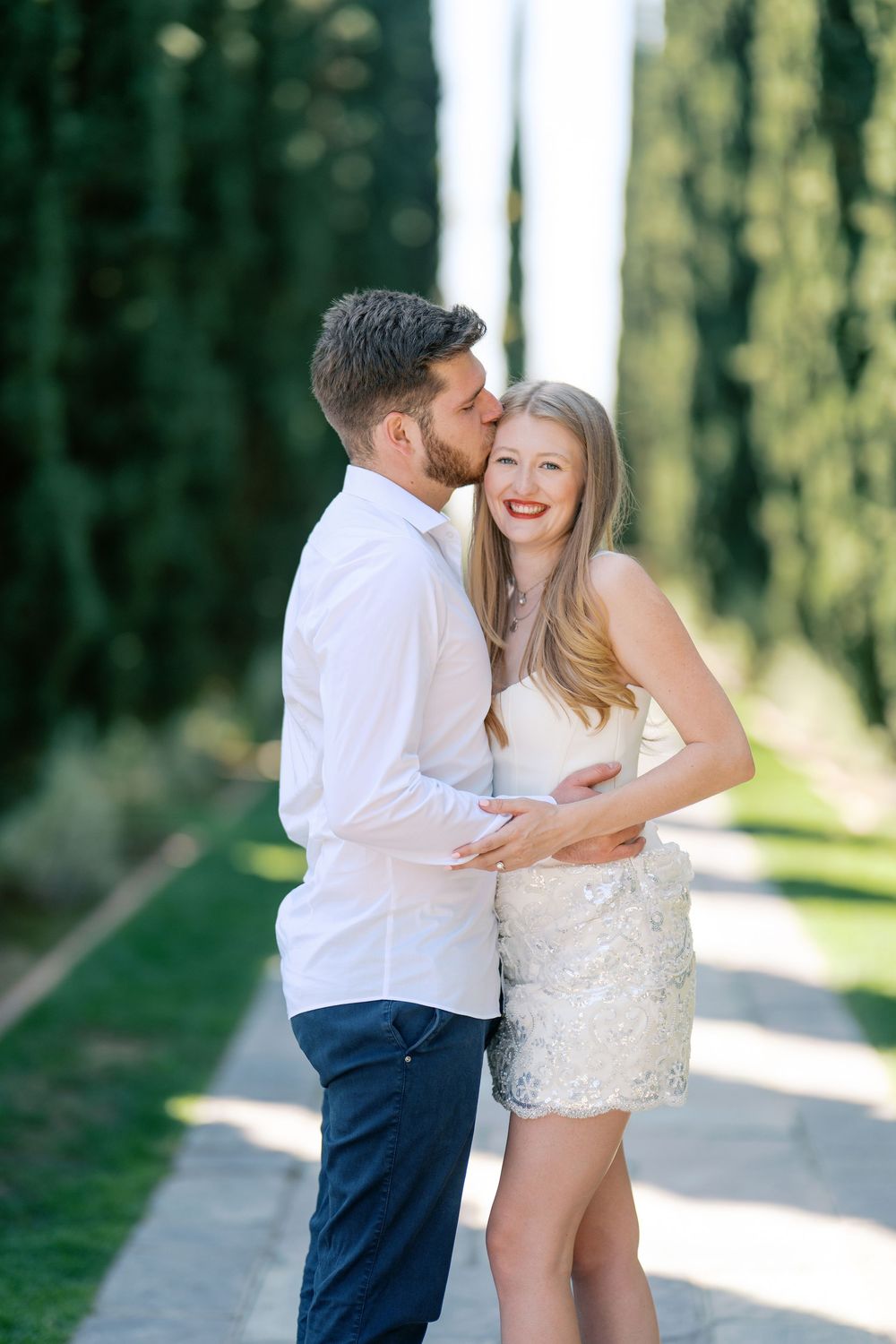 A couple shares a romantic kiss outdoors, with the woman smiling in a white dress amid lush green cypress trees.