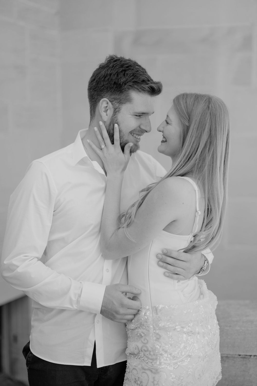 Smiling couple in white outfits sharing a romantic moment, woman touching mans beard during engagement photo session.