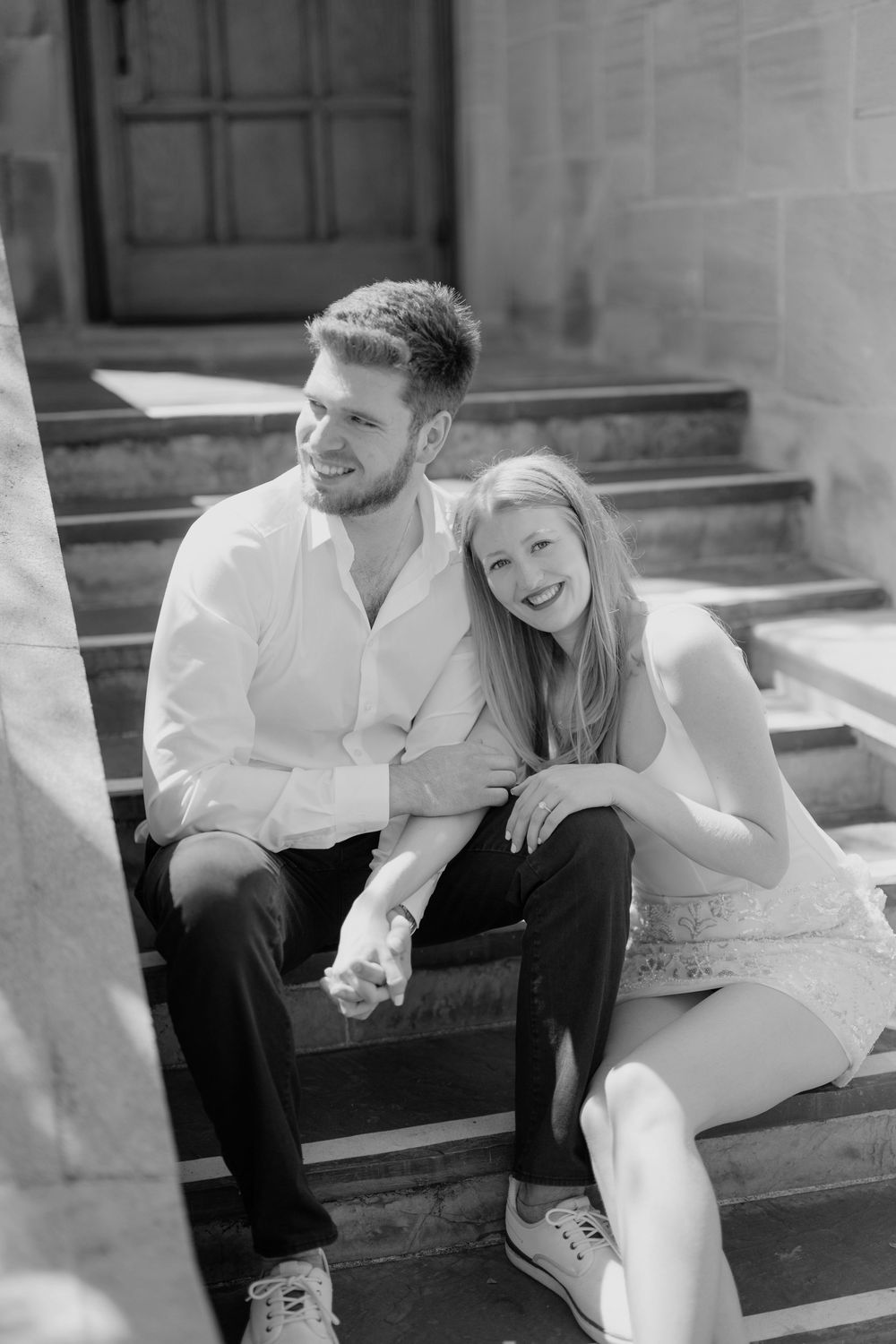 Black and white photo of a smiling couple sitting on stone steps, the woman laughing joyfully while leaning against the man.
