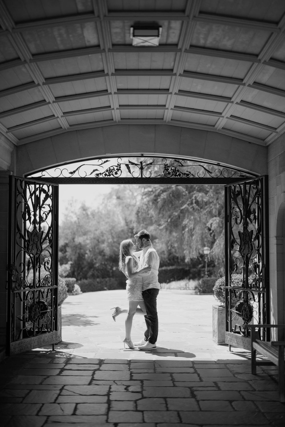 Black and white photo of a couple sharing a romantic moment framed by ornate iron gates.