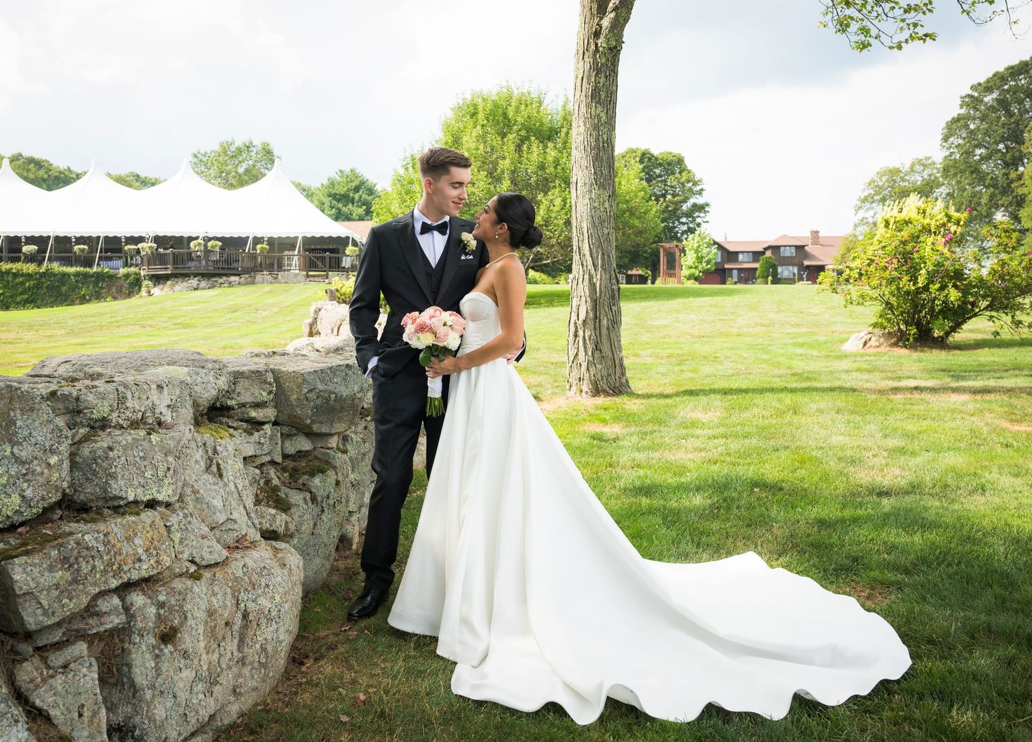 A smiling bride in a white gown and groom in a black tuxedo hold hands walking through a lush garden venue.