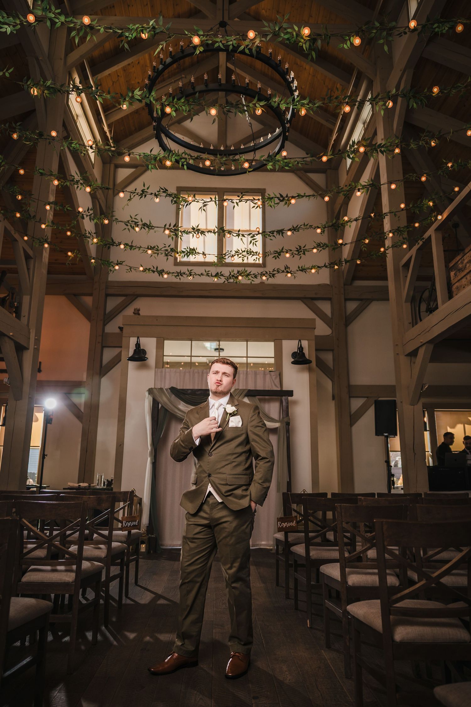 Groom portrait under chandelier and greenery at Briar Barn Inn ceremony space with rustic wood beams and string lights