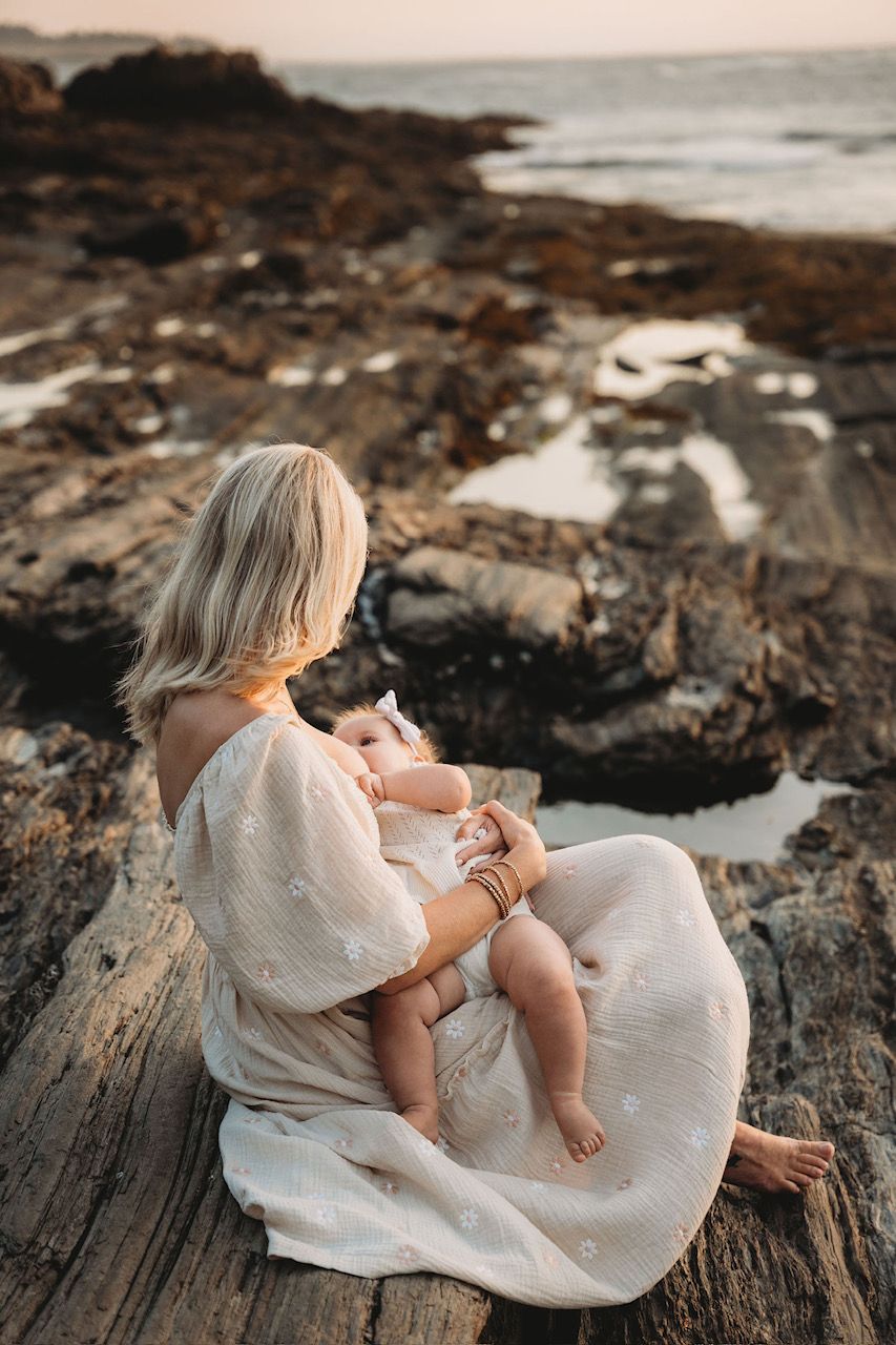 Mother in white dress breastfeeding baby on rocky coastal shoreline at sunset.