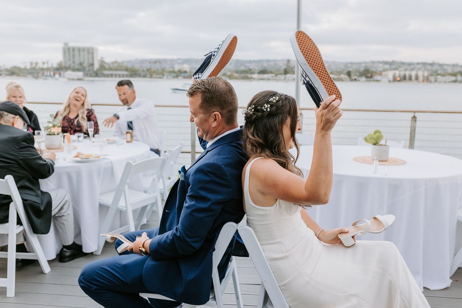 Bride and groom playing a wedding shoe game on a waterfront venue deck with guests seated at white tables at Tower Beach Club.