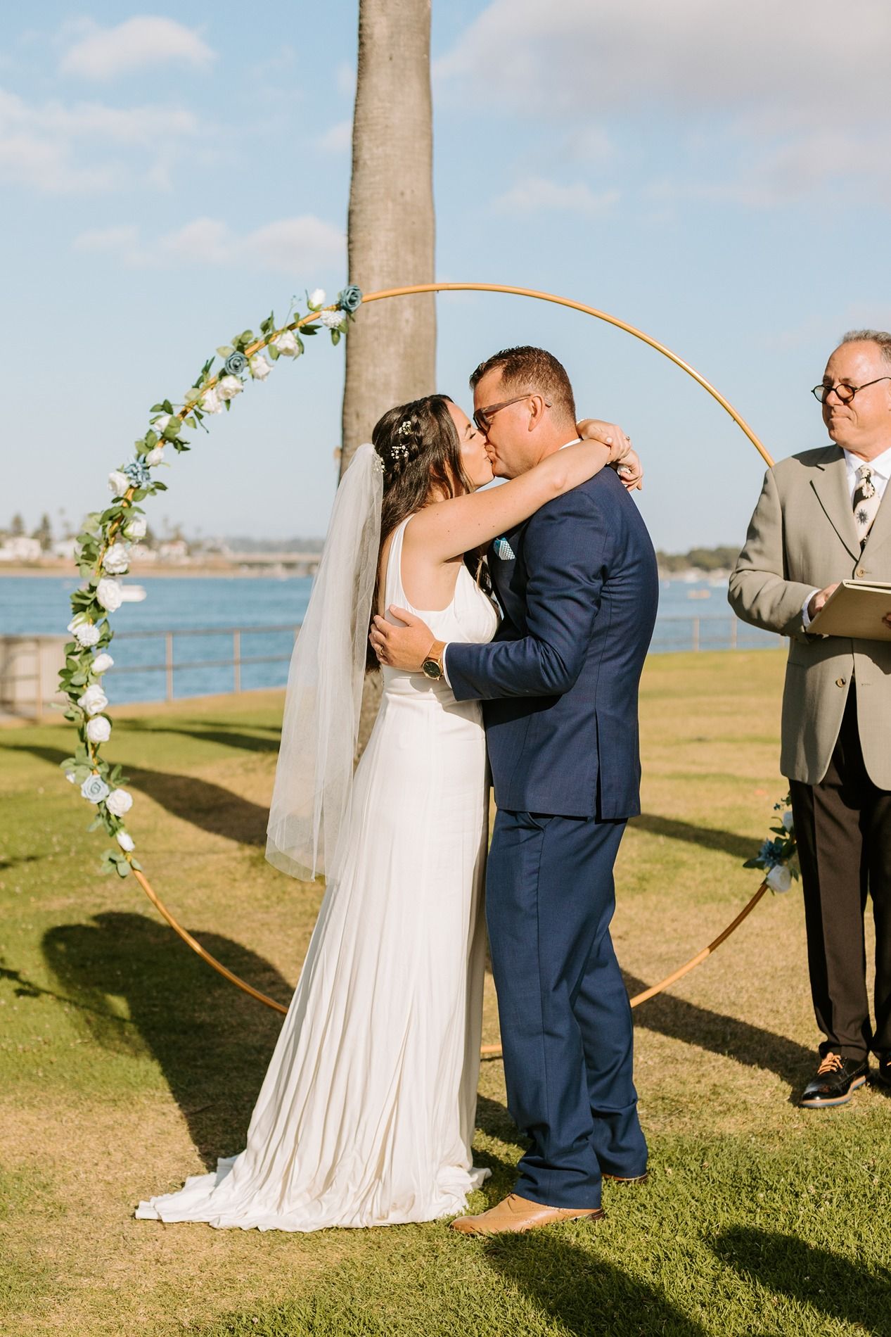 Bride and groom share first kiss at outdoor waterfront wedding ceremony under a circular floral arch at Tower Beach Club.