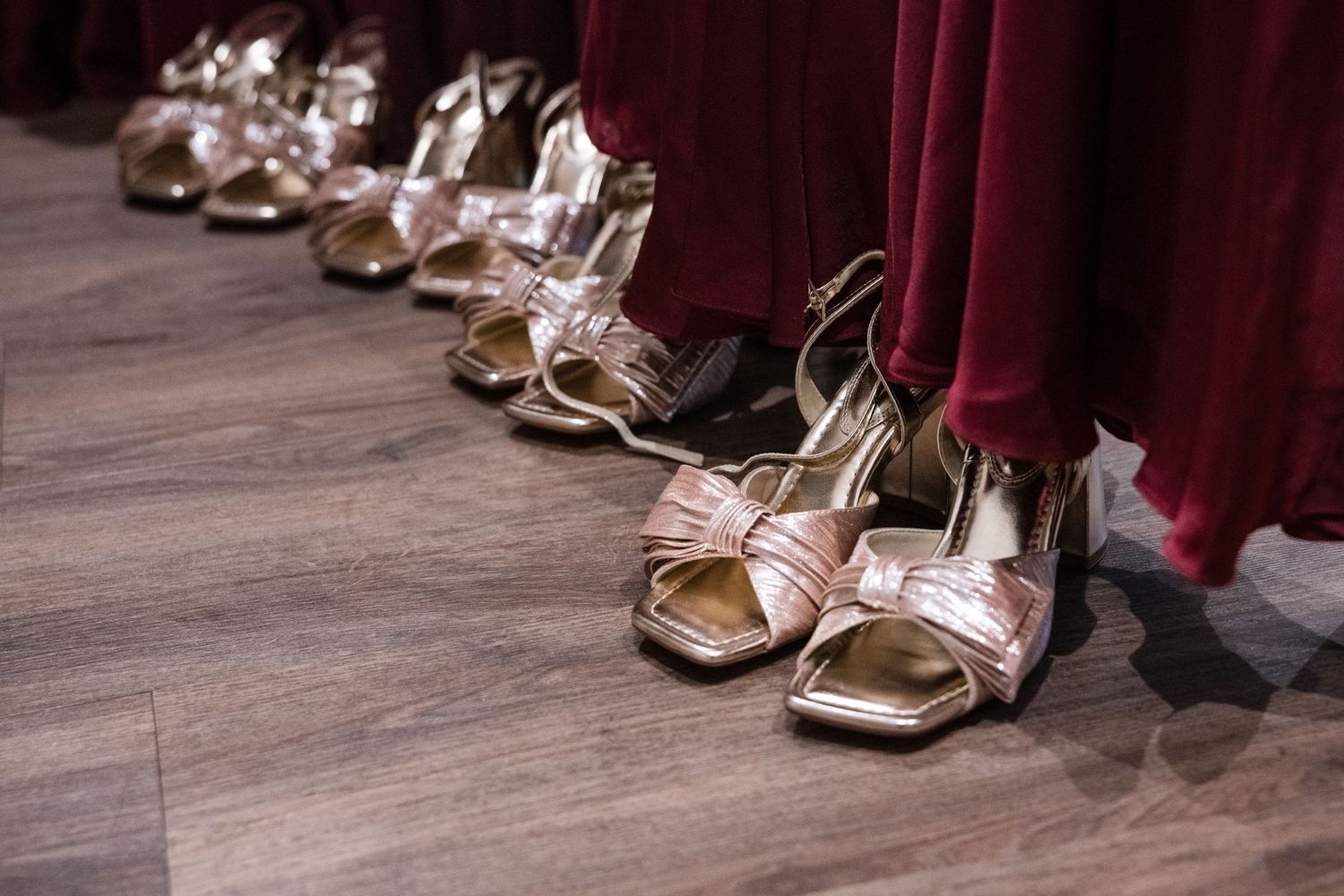 Row of gold strappy heels worn by bridesmaids in burgundy dresses on wooden floor.