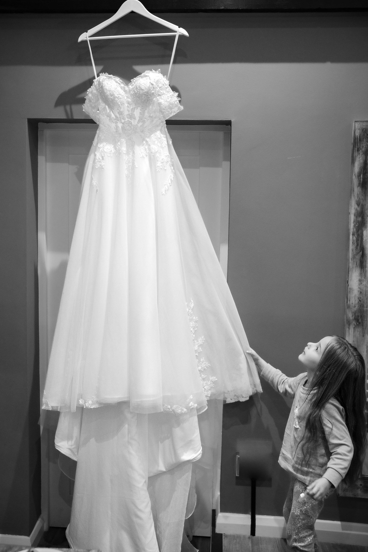 Black and white photo of young girl reaching up to touch elegant lace wedding dress on hanger.