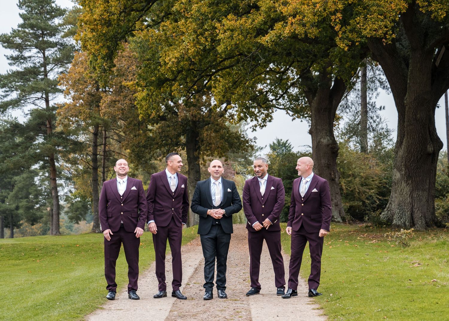 Groom in black tuxedo poses with four groomsmen in burgundy suits on tree-lined path at autumn wedding venue.