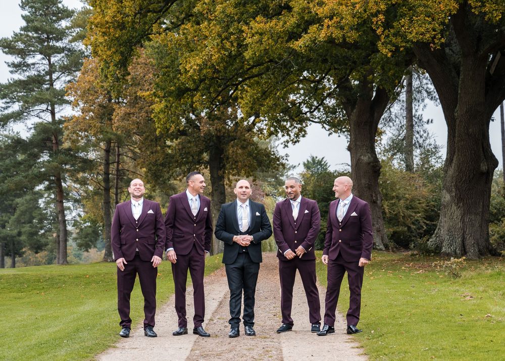 Groom in black tuxedo poses with four groomsmen in burgundy suits on tree-lined path at autumn wedding venue.