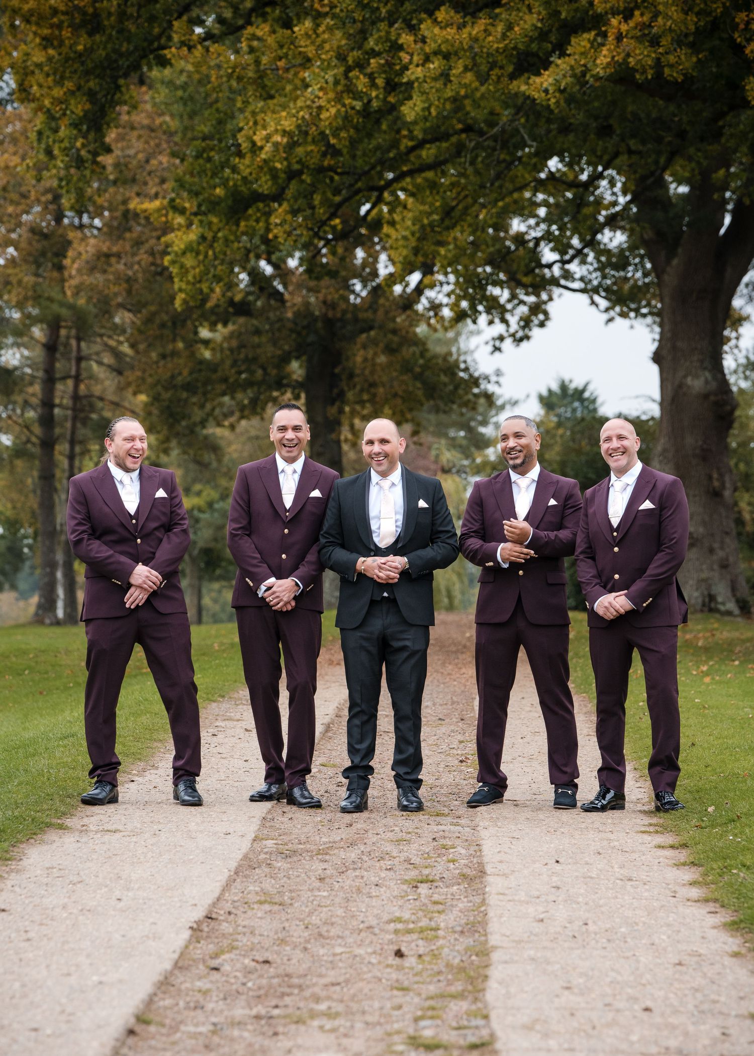Smiling groom in black suit stands with four groomsmen wearing matching burgundy three-piece suits on outdoor garden path.