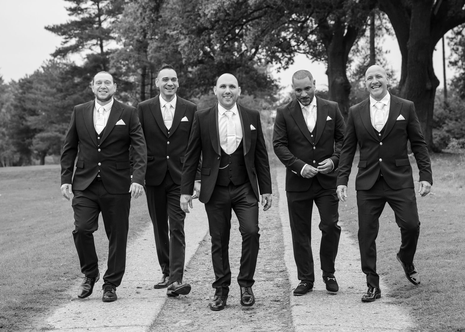 Black and white photo of groom and four groomsmen in suits walking confidently along a tree-lined path at wedding.