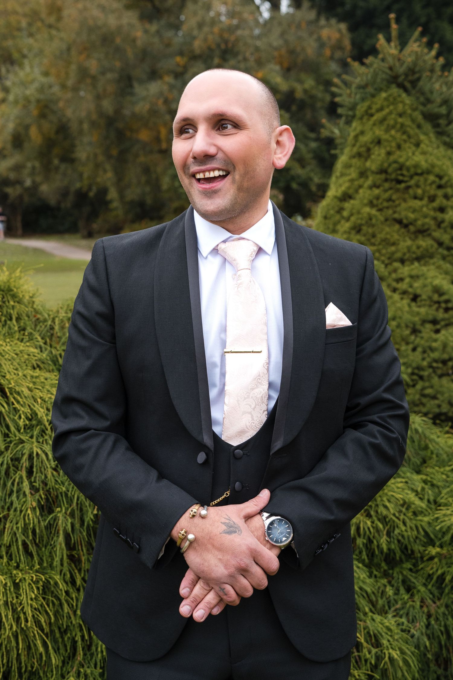 Smiling bald groom in black tuxedo with floral tie and pocket square poses outdoors among green hedges on wedding day.