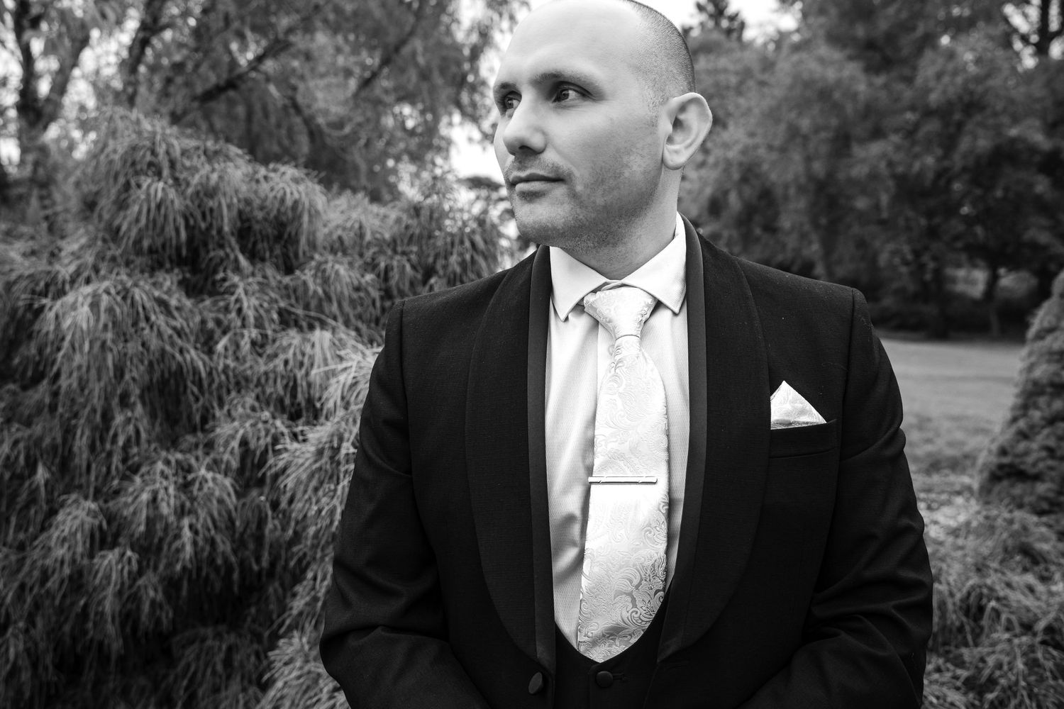 Black and white portrait of bald groom in black tuxedo with silk tie and pocket square standing near garden hedges.
