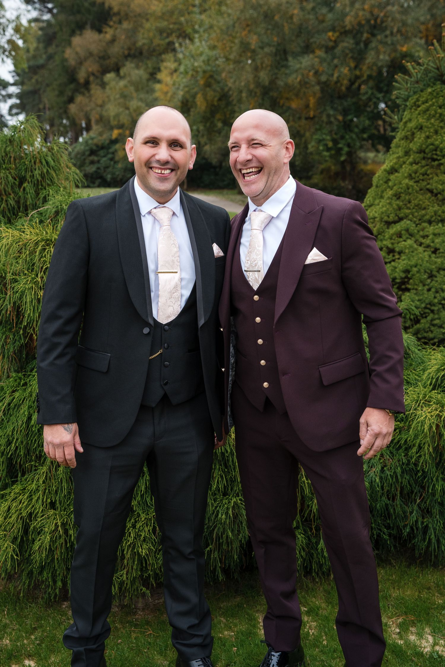 Groom in black tuxedo and best man in burgundy suit laugh together outdoors among green hedges at autumn wedding.
