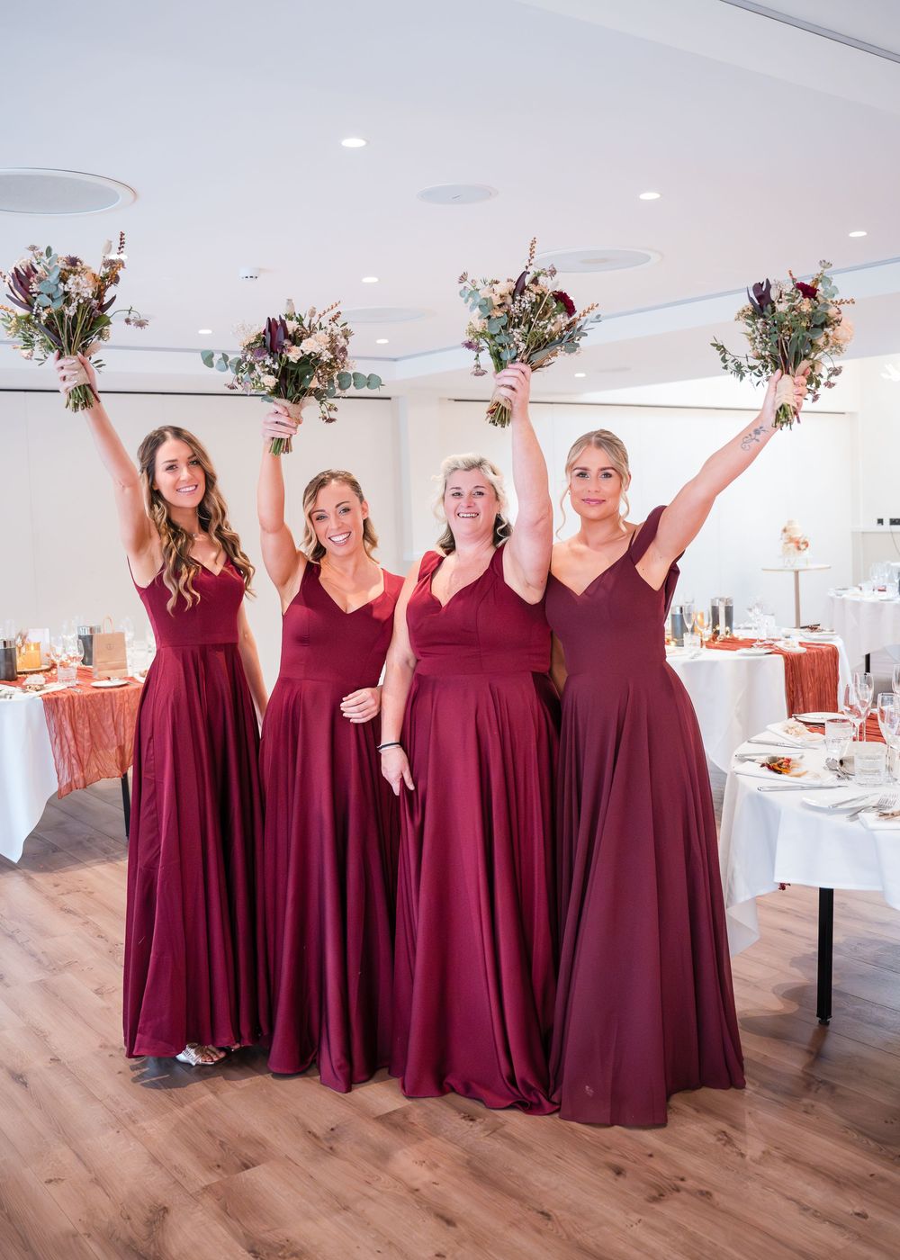 Four bridesmaids in floor-length burgundy gowns joyfully raise bouquets of wildflowers inside elegant wedding reception venue.