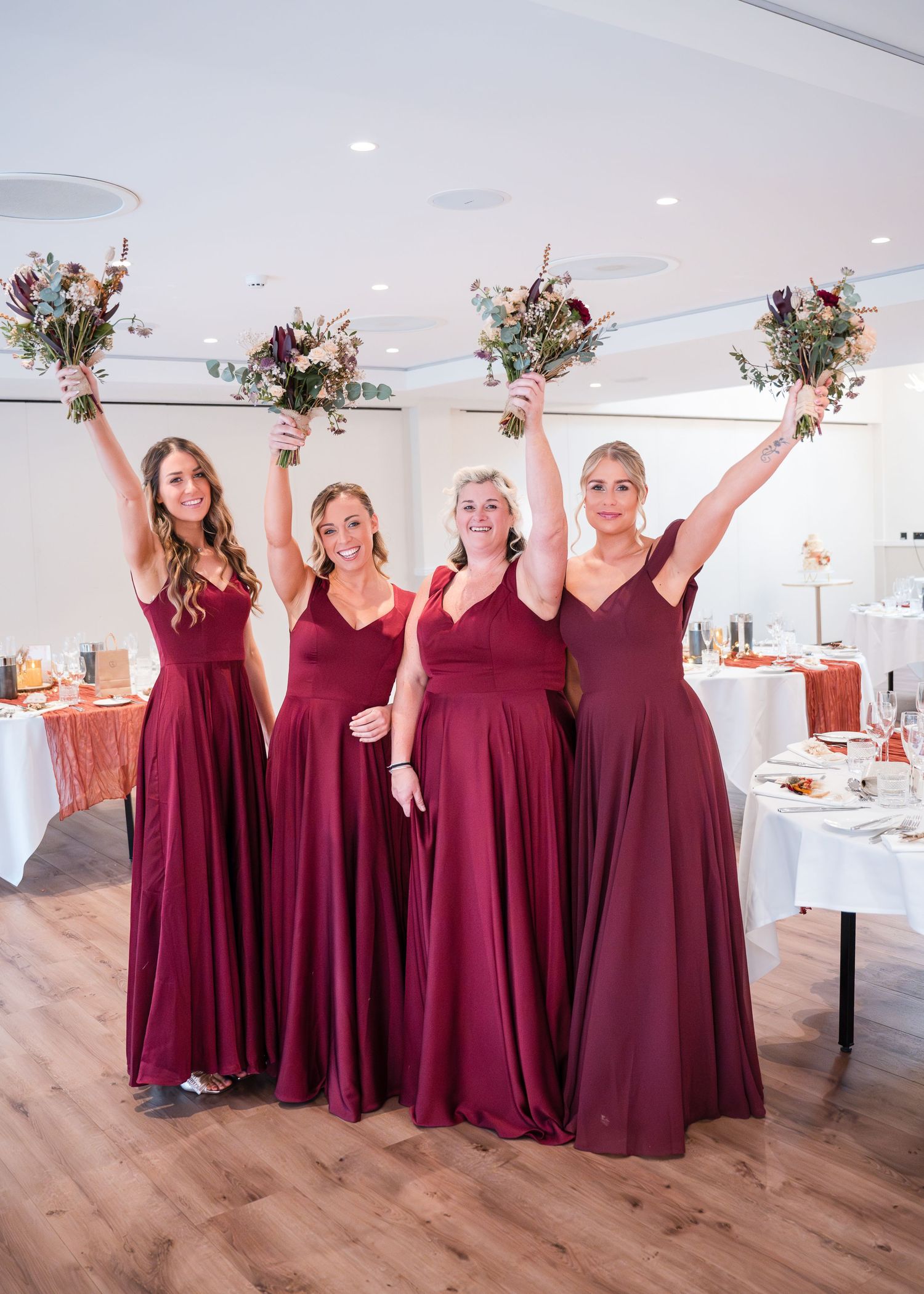 Four bridesmaids in floor-length burgundy gowns joyfully raise bouquets of wildflowers inside elegant wedding reception venue.