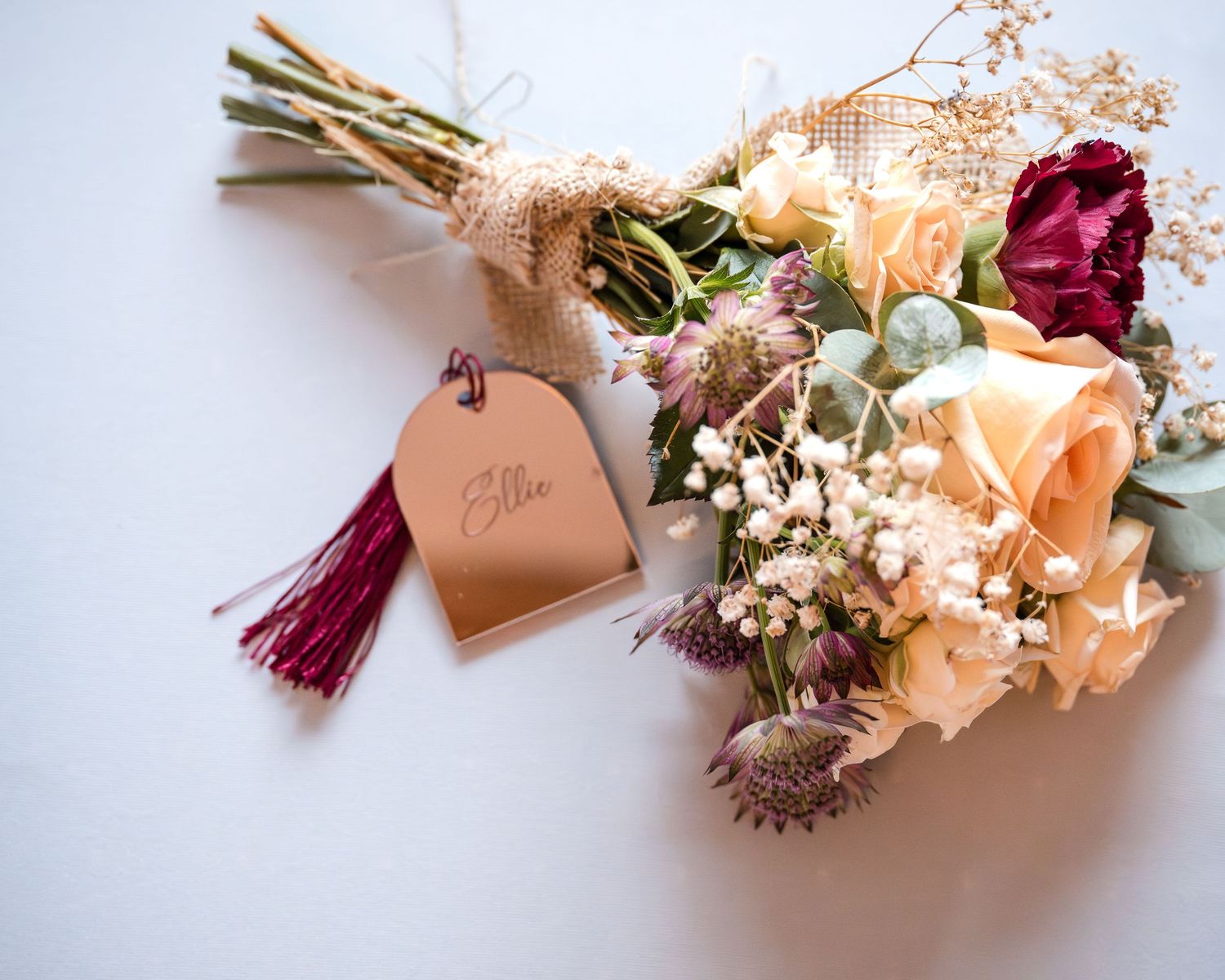 Elegant wedding bouquet with peach roses, burgundy flowers, and a personalized leather tag on a white background.