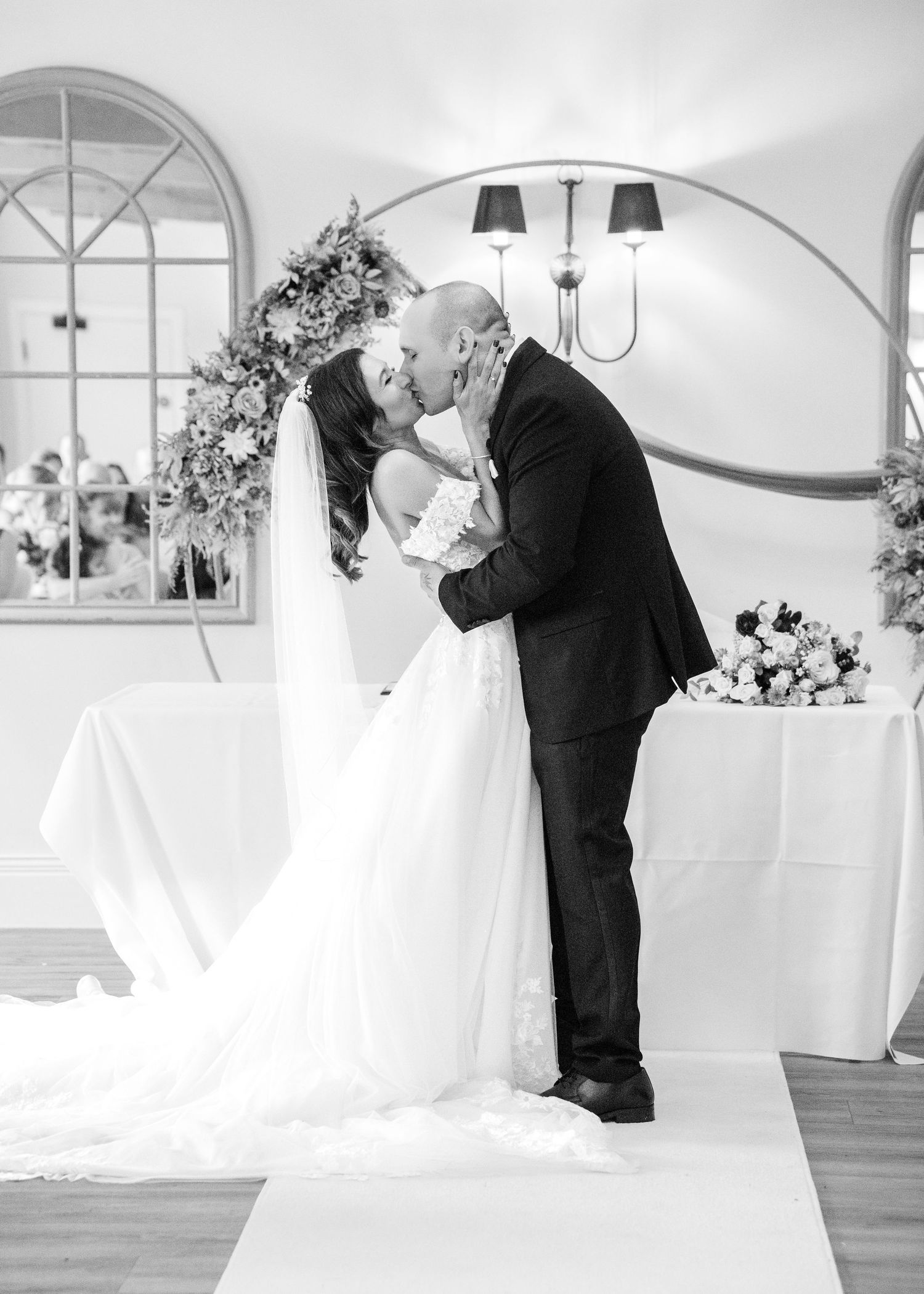 Romantic black and white photo of newlyweds kissing at the altar surrounded by floral arrangements.