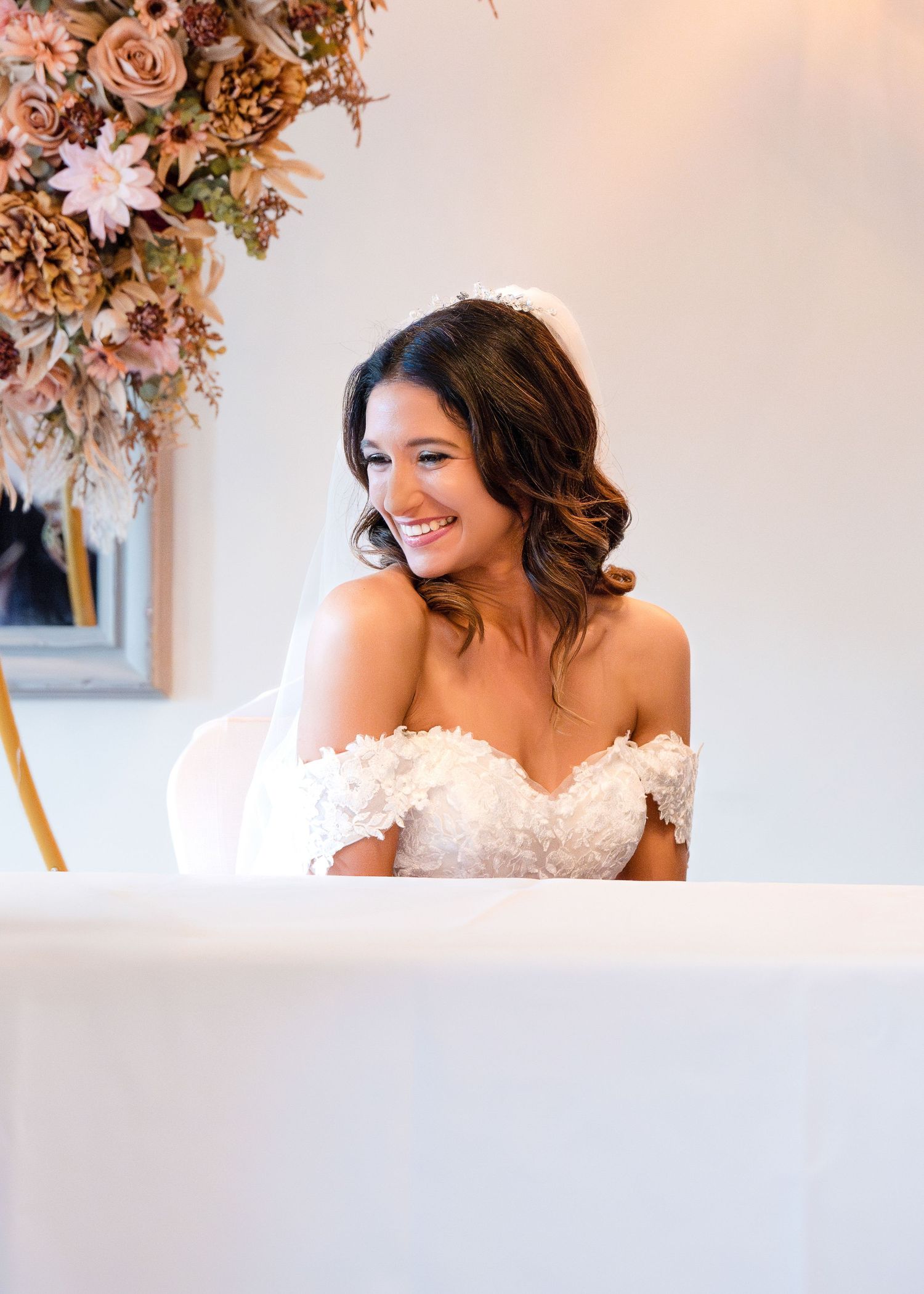 Smiling bride in an off-shoulder lace wedding dress seated at the signing table with floral backdrop.