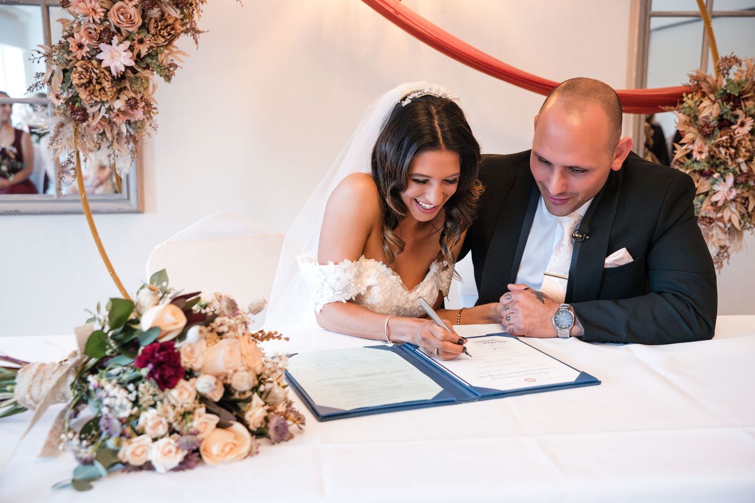 Bride and groom signing their wedding certificate together at a white table with a bridal bouquet nearby.