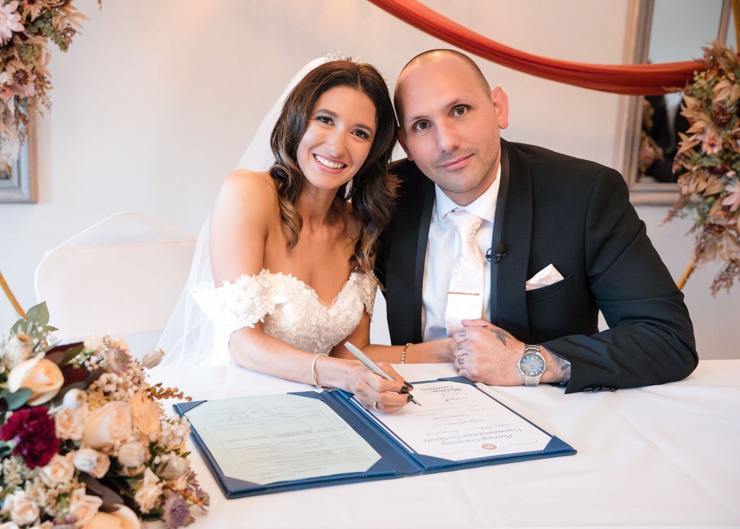Newlyweds smile while signing the wedding register at a flower-adorned table during their ceremony.
