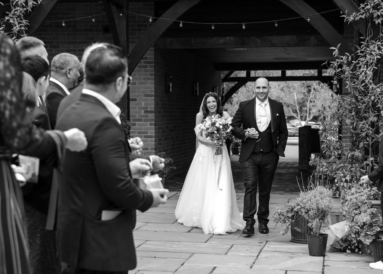 Black and white photo of bride and groom emerging from a rustic venue entrance to cheering wedding guests.