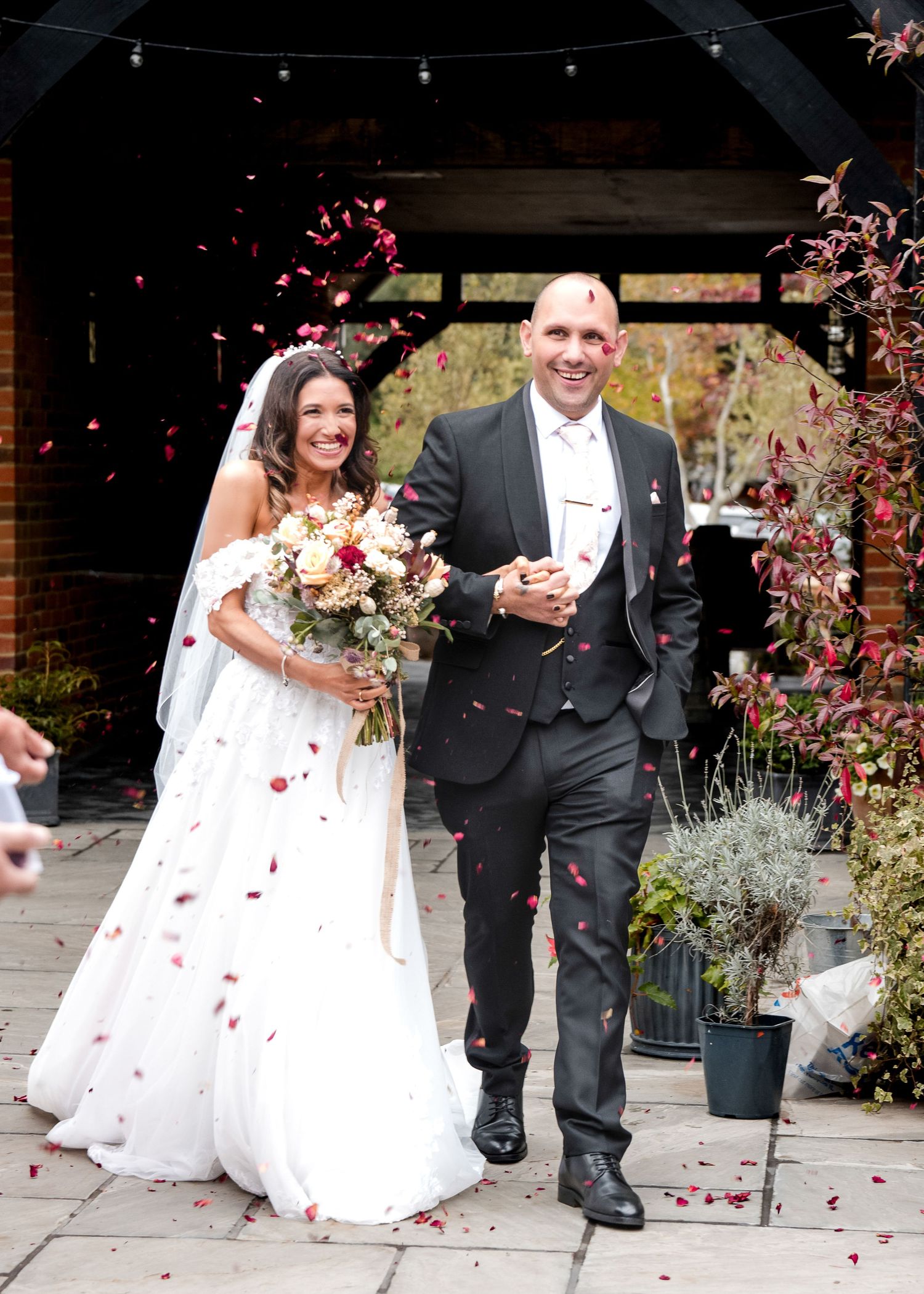 Joyful newlyweds walk through a shower of red confetti petals outside a rustic wedding venue.