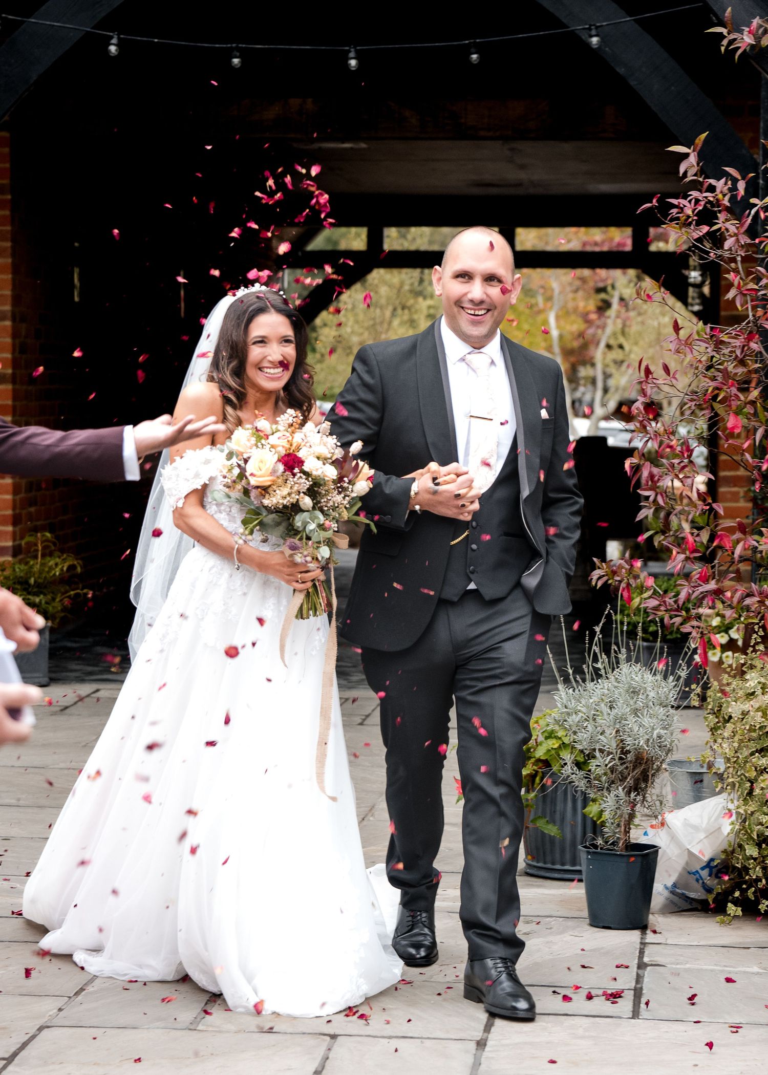 Bride and groom walk through confetti shower outside rustic wedding venue, bride holding autumn bouquet.
