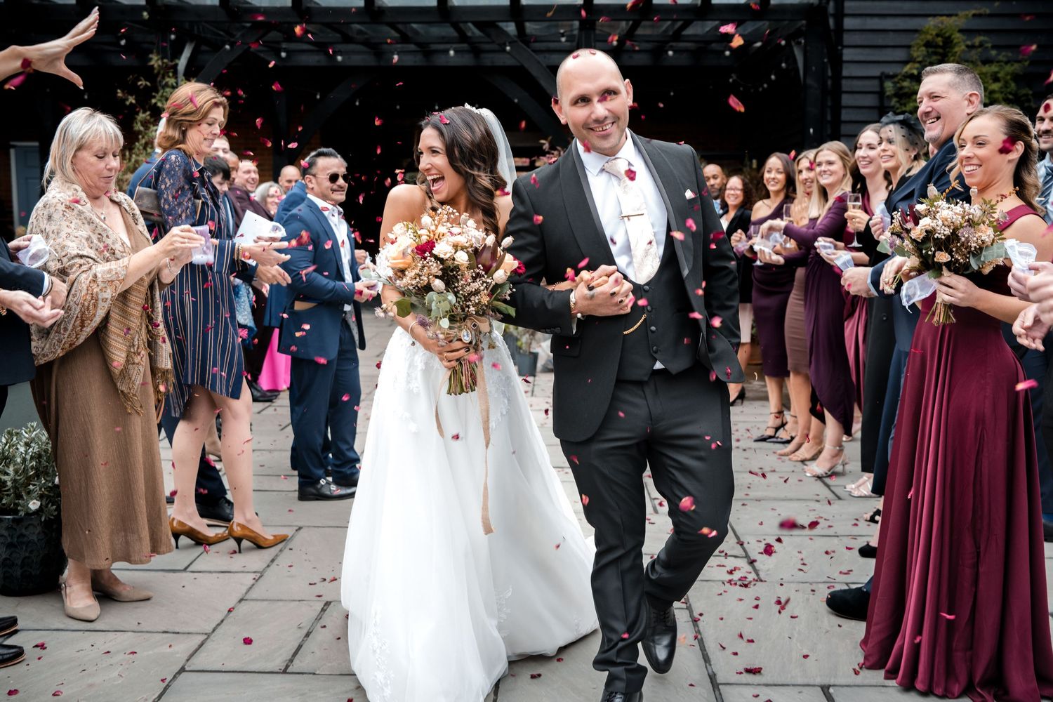 Smiling bride and groom exit wedding venue as guests throw red and gold confetti petals.