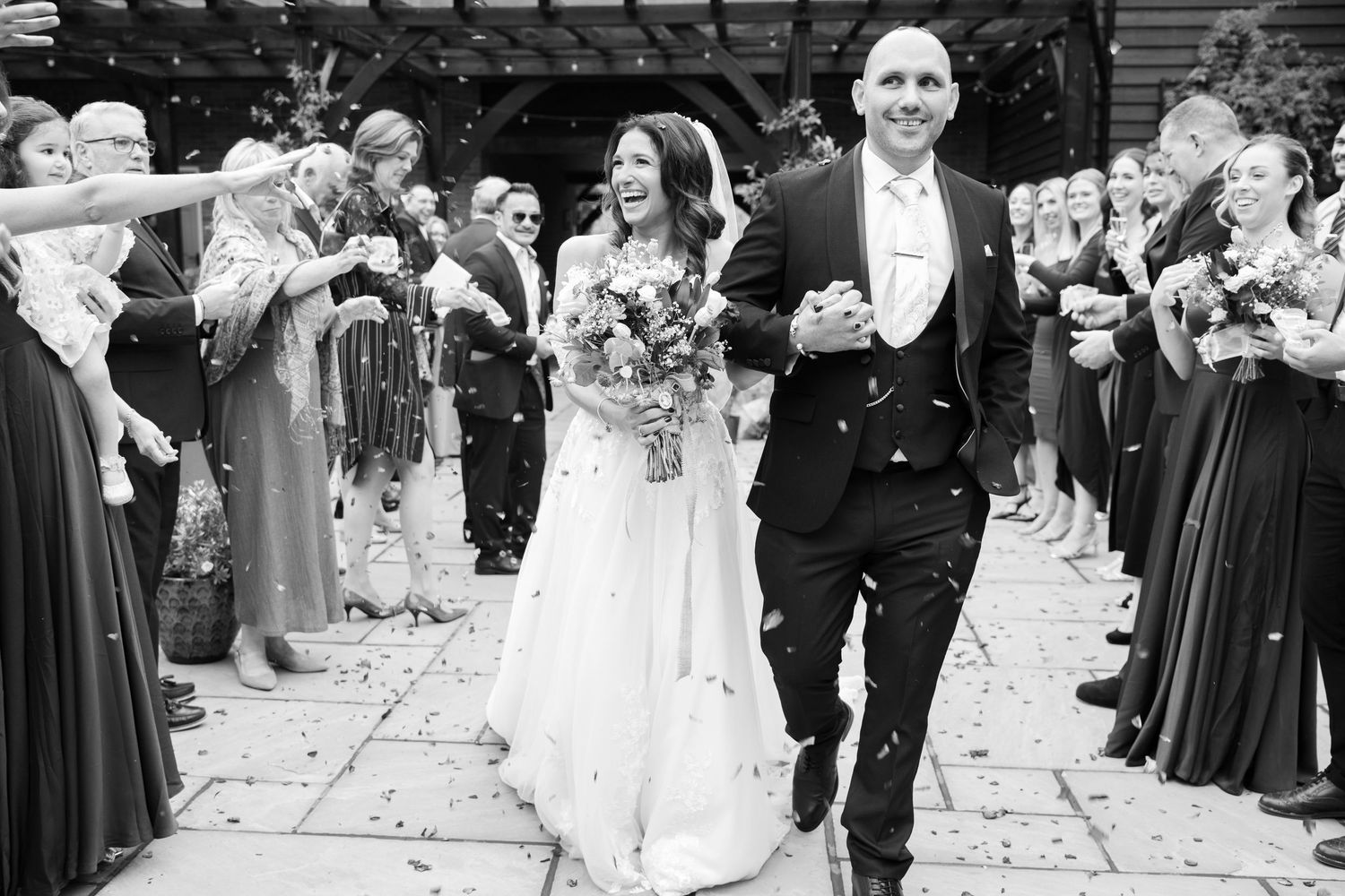 Black and white photo of newlyweds walking through confetti tunnel created by wedding guests.