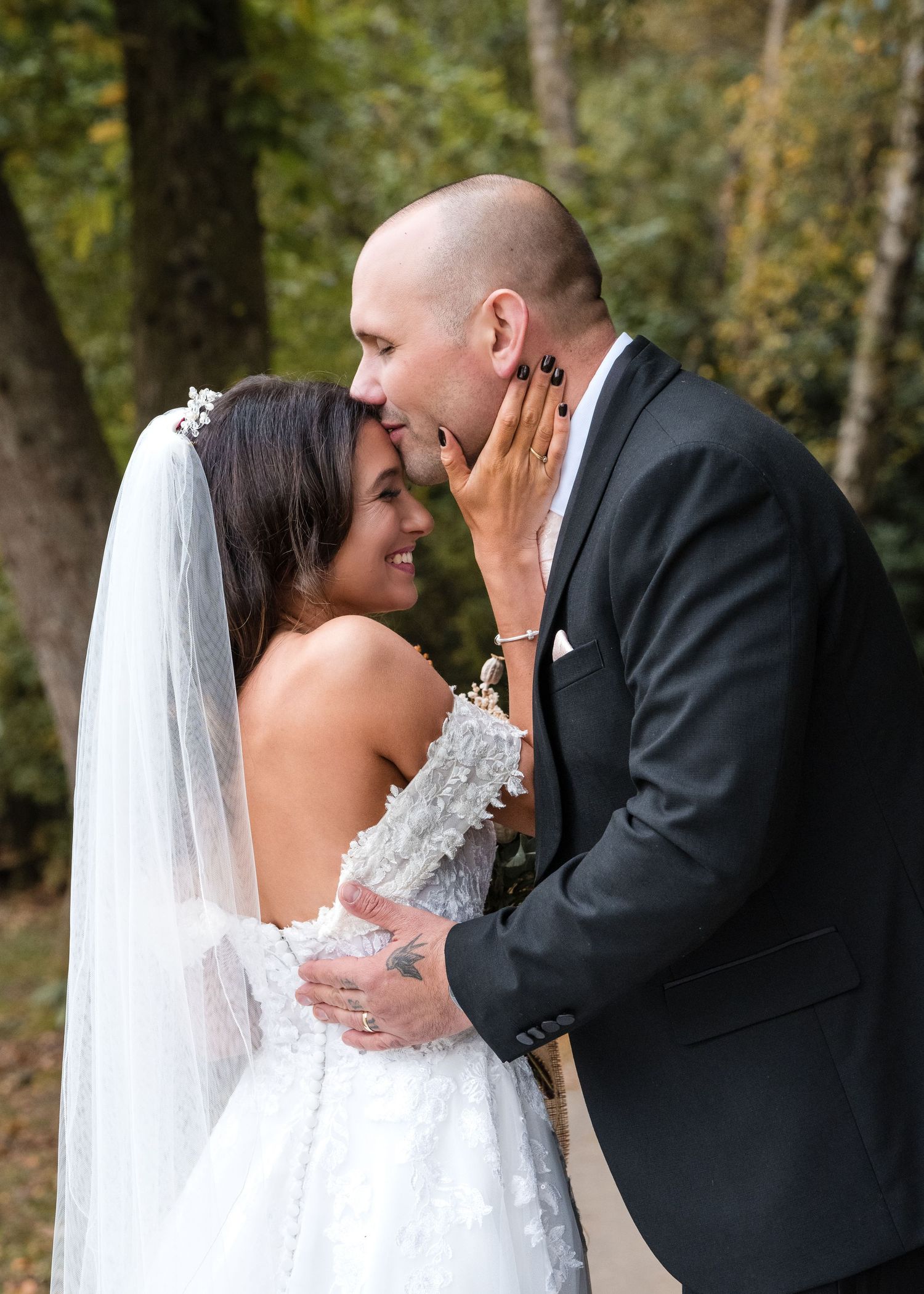 Bride smiles as groom tenderly cups her face for a kiss in an autumn woodland setting.