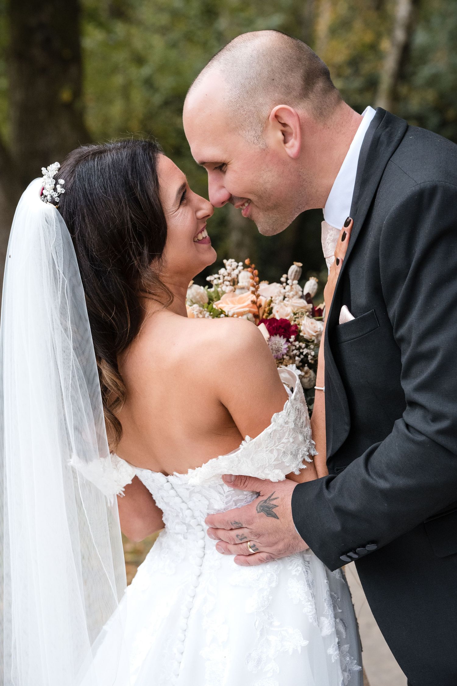 Bride and groom share a romantic nose-to-nose moment holding an autumn wedding bouquet outdoors.