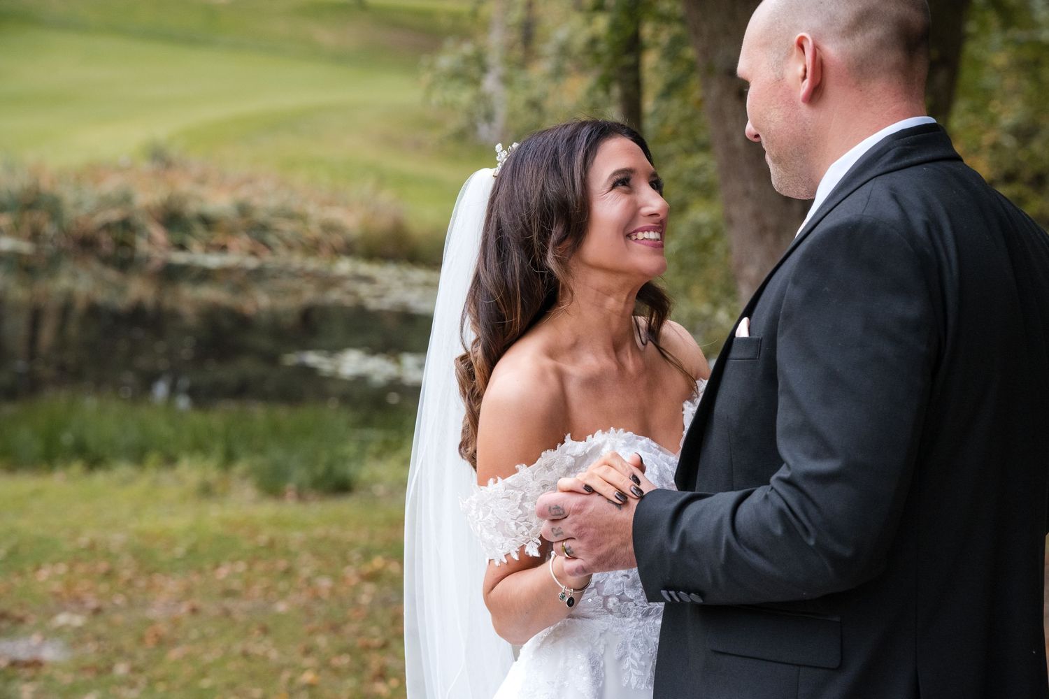 Radiant bride laughs joyfully while holding hands with groom near a scenic outdoor water feature.