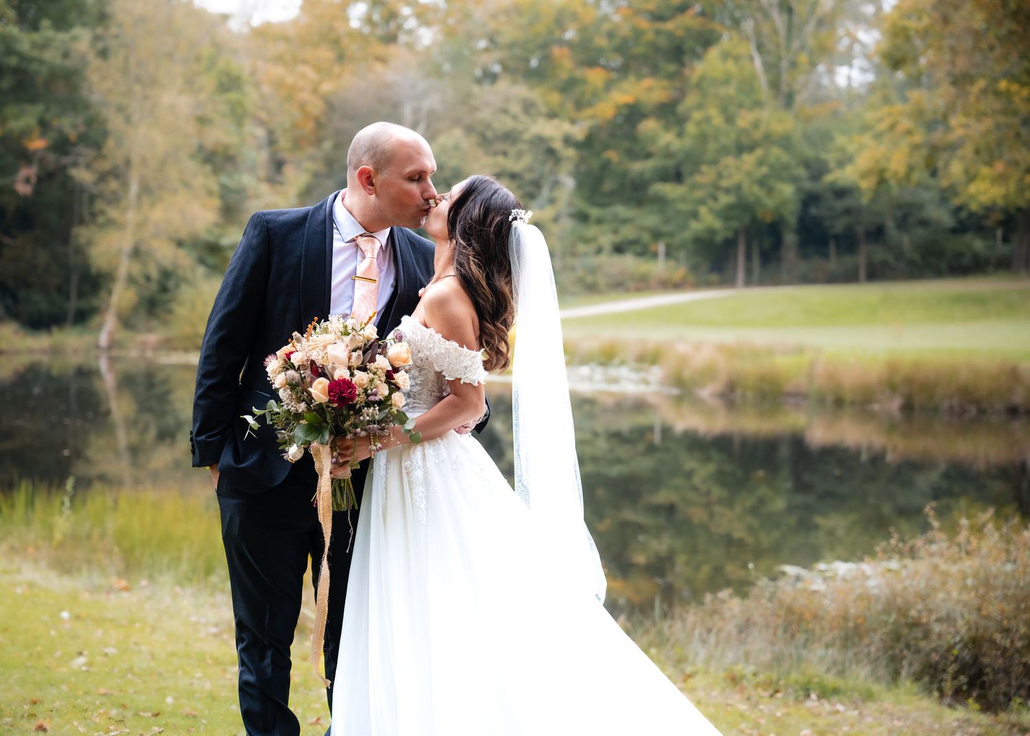 Bride and groom kissing near a serene pond surrounded by autumn foliage on their wedding day.