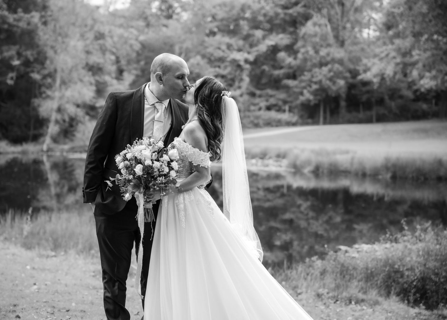 Black and white photo of bride and groom sharing a romantic kiss by a tranquil pond.